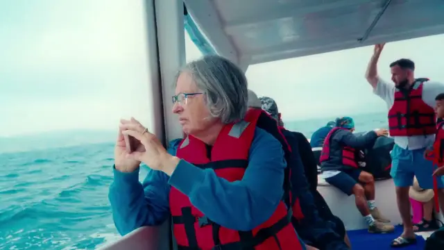Video still: a traveller in a life jacket photographing the sea from a boat